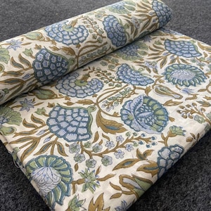May include: Rolled and flat floral fabric featuring a repeating pattern of blue, green, and brown flowers and foliage on an off-white background. The fabric is folded and rolled, showing the detailed design.