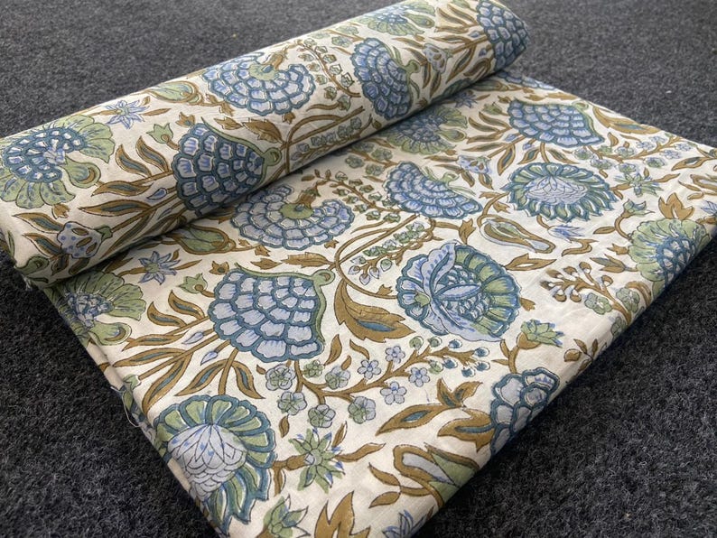 May include: Rolled and flat floral fabric featuring a repeating pattern of blue, green, and brown flowers and foliage on an off-white background. The fabric is folded and rolled, showing the detailed design.