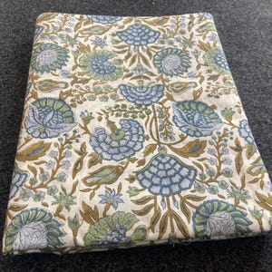 May include: Folded fabric with a floral pattern in shades of blue, green, and brown on a cream background. The design features stylized flowers and foliage. The fabric is likely cotton or linen.