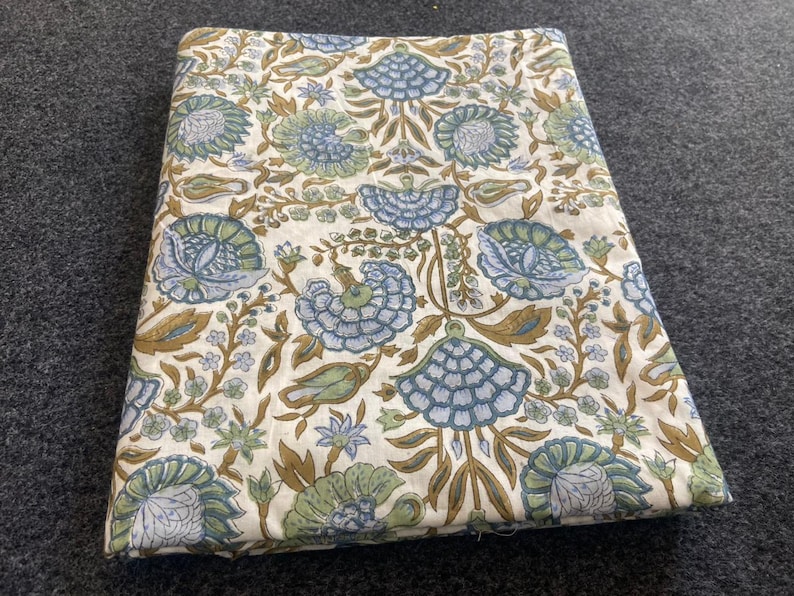 May include: Folded fabric with a floral pattern in shades of blue, green, and brown on a cream background. The design features stylized flowers and foliage. The fabric is likely cotton or linen.