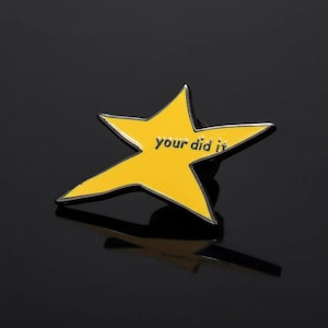 Könnte beinhalten: Ein gelber sternförmiger Emaille-Anstecker mit dem Text "Your did it" in Schwarz. Der Anstecker hat einen schwarzen Rand und einen Nadelverschluss. Der Stern ist vor einem dunklen Hintergrund platziert.