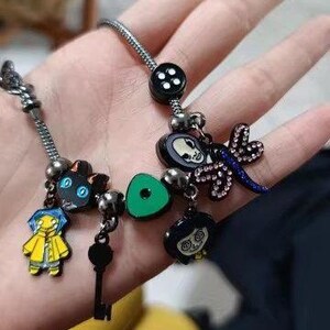 Puede incluir: Una pulsera de plata con varios abalorios de esmalte. Los abalorios incluyen un gato negro, una llave, un tri&aacute;ngulo verde, una lib&eacute;lula y personajes de una pel&iacute;cula. La pulsera se sostiene en una mano.