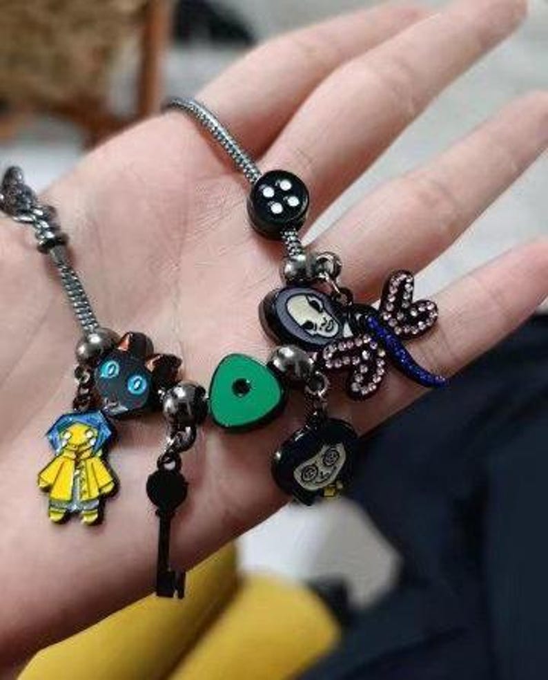 Puede incluir: Una pulsera de plata con varios abalorios de esmalte. Los abalorios incluyen un gato negro, una llave, un tri&aacute;ngulo verde, una lib&eacute;lula y personajes de una pel&iacute;cula. La pulsera se sostiene en una mano.
