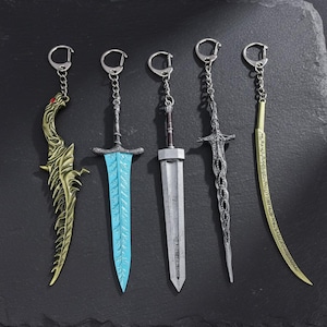 Op de afbeelding: Vijf miniatuur zwaard sleutelhangers met zilveren kettingen en sluitingen. De zwaarden variëren in ontwerp, waaronder een gouden draakachtige kling, een blauwlemmet zwaard, een zilveren breedzwaard, een gedetailleerd zilveren zwaard en een gebogen gouden kling.