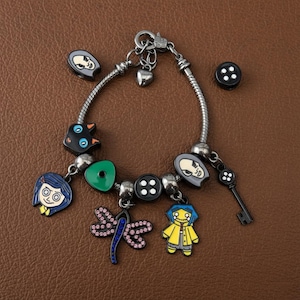 Puede incluir: Pulsera de abalorios plateada con varios abalorios de esmalte. Los abalorios incluyen una llave, botones, una lib&eacute;lula y colgantes tem&aacute;ticos de personajes en azul, amarillo y negro. La pulsera se muestra sobre una superficie marr&oacute;n.