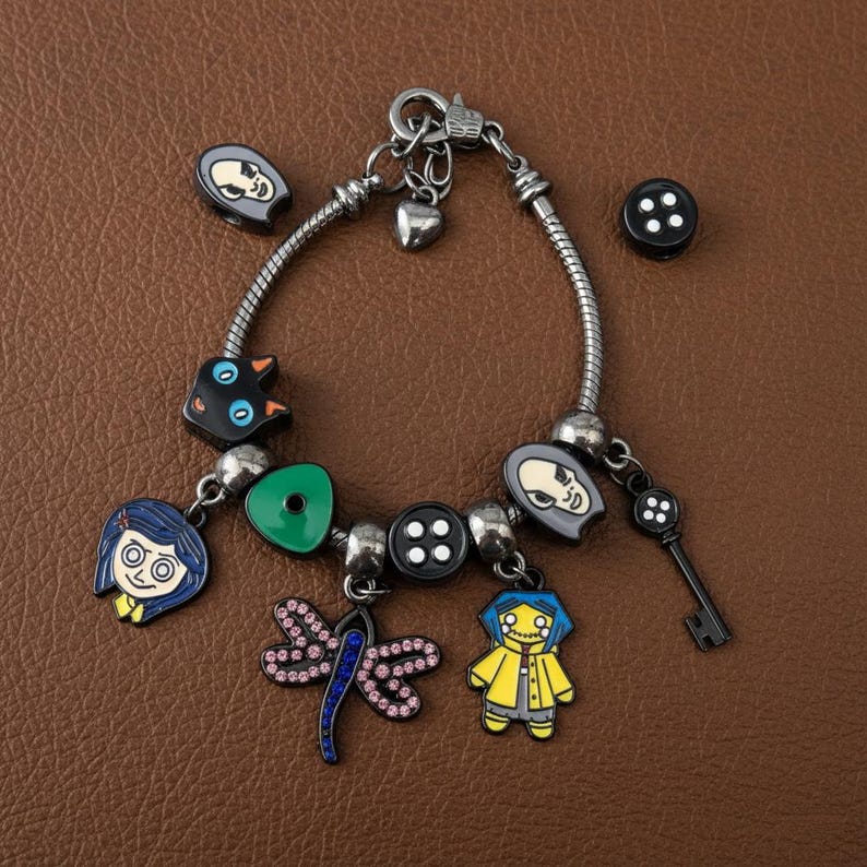 Puede incluir: Pulsera de abalorios plateada con varios abalorios de esmalte. Los abalorios incluyen una llave, botones, una lib&eacute;lula y colgantes tem&aacute;ticos de personajes en azul, amarillo y negro. La pulsera se muestra sobre una superficie marr&oacute;n.