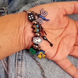 Puede incluir: Una pulsera de plata con varios dijes coloridos. Los dijes incluyen un bot&oacute;n negro, una lib&eacute;lula, una llave y dijes de personajes de dibujos animados. La pulsera tiene una cadena con un dije en forma de coraz&oacute;n.