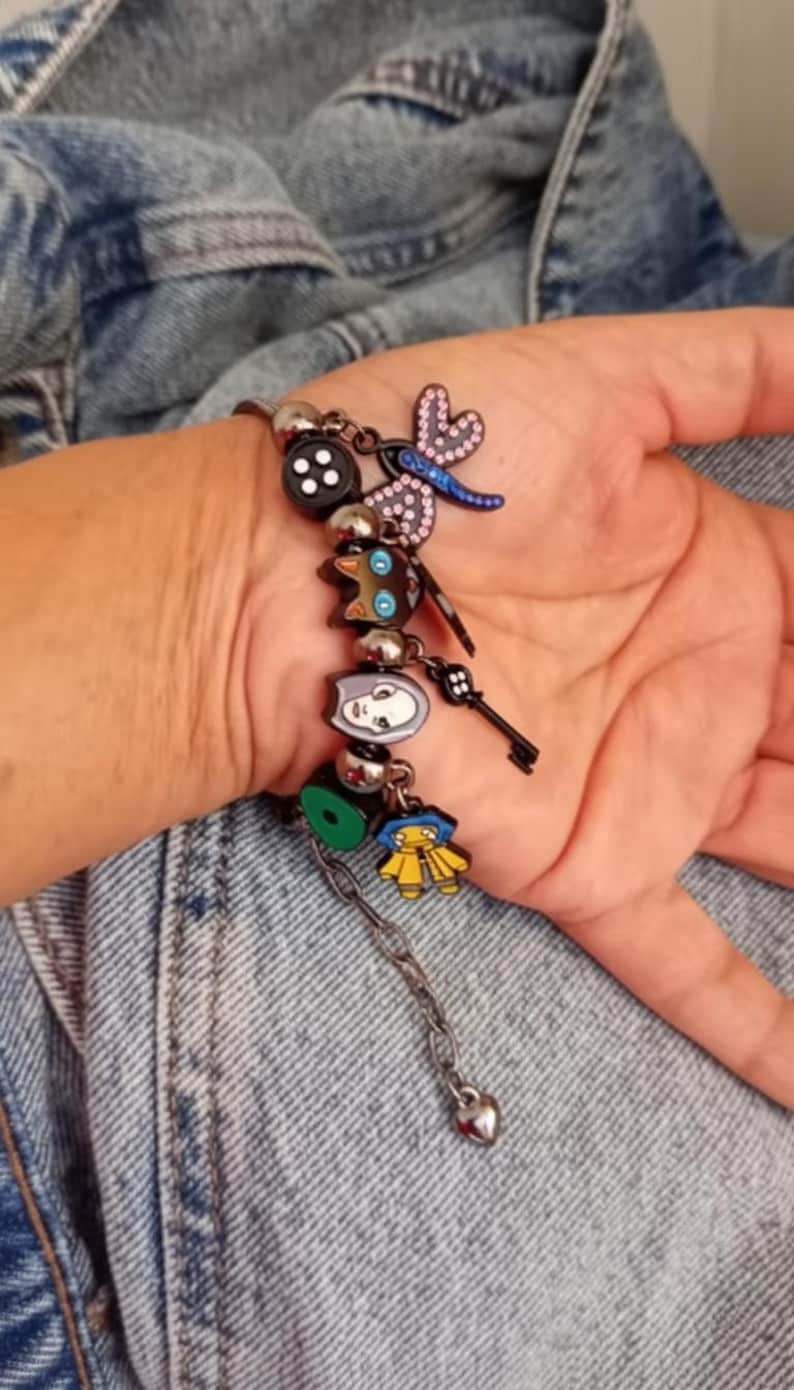 Puede incluir: Una pulsera de plata con varios dijes coloridos. Los dijes incluyen un bot&oacute;n negro, una lib&eacute;lula, una llave y dijes de personajes de dibujos animados. La pulsera tiene una cadena con un dije en forma de coraz&oacute;n.
