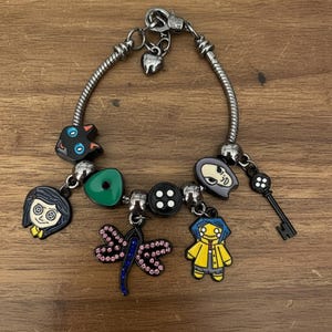 Puede incluir: Pulsera de abalorios plateada con varios abalorios de esmalte. Los abalorios incluyen un gato negro, un coraz&oacute;n verde, una lib&eacute;lula, una llave y personajes con abrigos amarillos. La pulsera est&aacute; sobre una superficie de madera.
