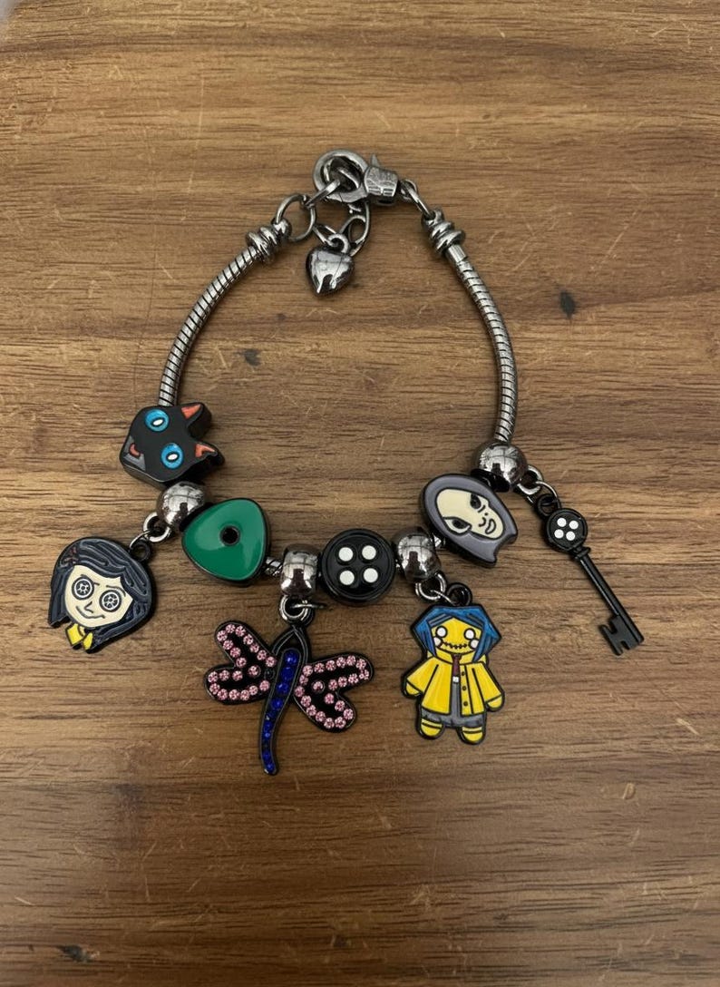 Puede incluir: Pulsera de abalorios plateada con varios abalorios de esmalte. Los abalorios incluyen un gato negro, un coraz&oacute;n verde, una lib&eacute;lula, una llave y personajes con abrigos amarillos. La pulsera est&aacute; sobre una superficie de madera.