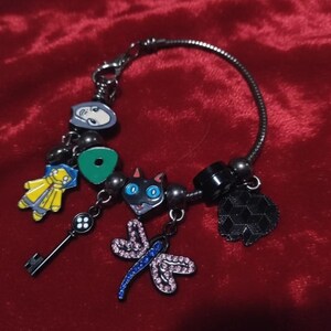 Puede incluir: Una pulsera de plata con varios abalorios de colores. Los abalorios incluyen una llave, una figura amarilla, un gato negro con ojos azules, una lib&eacute;lula y un abalorio negro en forma de coraz&oacute;n. La pulsera est&aacute; sobre una superficie de terciopelo rojo.