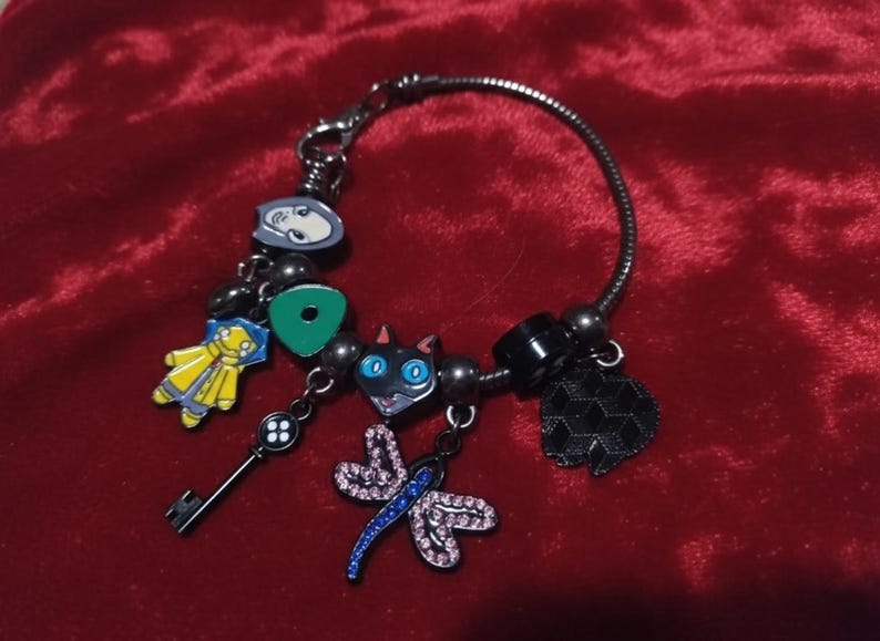 Puede incluir: Una pulsera de plata con varios abalorios de colores. Los abalorios incluyen una llave, una figura amarilla, un gato negro con ojos azules, una lib&eacute;lula y un abalorio negro en forma de coraz&oacute;n. La pulsera est&aacute; sobre una superficie de terciopelo rojo.
