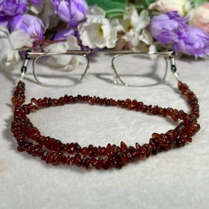 May include: A pair of clear-framed eyeglasses attached to a long beaded chain made of small, irregular, deep reddish-brown amber-colored stone chips. The chain features metallic silver-toned connectors at each end that secure to the eyeglass temples.