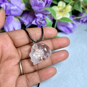 May include: A clear quartz crystal pendant carved in the shape of a skull, suspended from a black cord necklace. The pendant is held in a hand, with a blurred background of purple flowers.