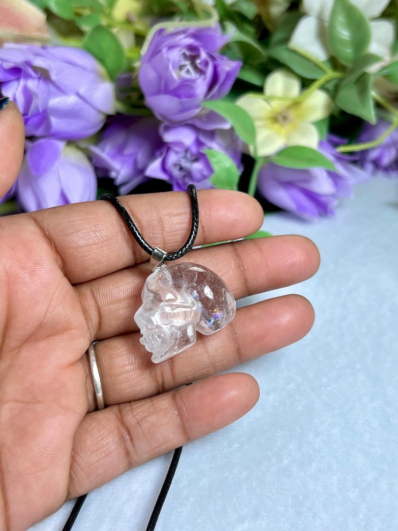 May include: A clear quartz crystal pendant carved in the shape of a skull, suspended from a black cord necklace. The pendant is held in a hand, with a blurred background of purple flowers.