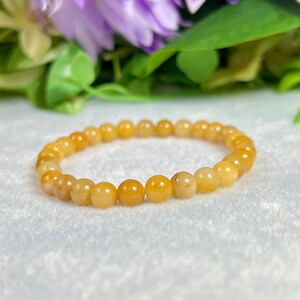 May include: A golden yellow beaded bracelet. The bracelet is made of small, round beads with a polished finish. The beads are strung together on an elastic band. The background features purple flowers and green leaves.