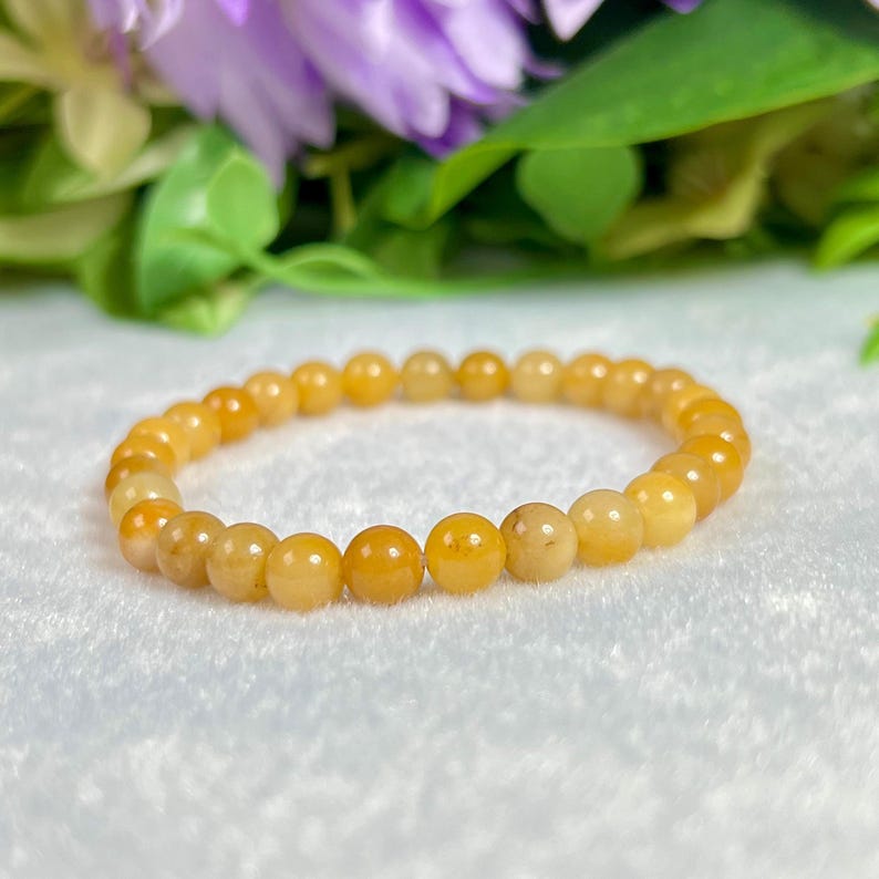 May include: A golden yellow beaded bracelet. The bracelet is made of small, round beads with a polished finish. The beads are strung together on an elastic band. The background features purple flowers and green leaves.