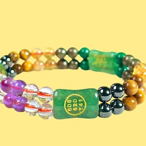 K&ouml;nnte beinhalten: Doppelreihiges Armband mit einer Mischung aus farbigen Perlen, darunter klare, lila, braune und gr&uuml;ne Steine. Das Armband hat zwei gr&uuml;ne rechteckige Jadest&uuml;cke mit goldenem Text. Der Hintergrund ist einfarbig gelb.