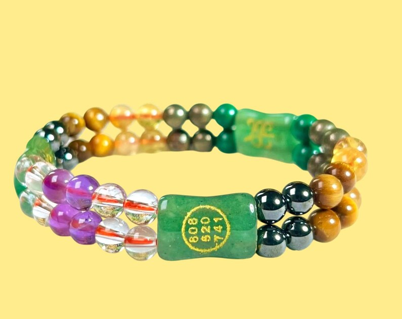 K&ouml;nnte beinhalten: Doppelreihiges Armband mit einer Mischung aus farbigen Perlen, darunter klare, lila, braune und gr&uuml;ne Steine. Das Armband hat zwei gr&uuml;ne rechteckige Jadest&uuml;cke mit goldenem Text. Der Hintergrund ist einfarbig gelb.