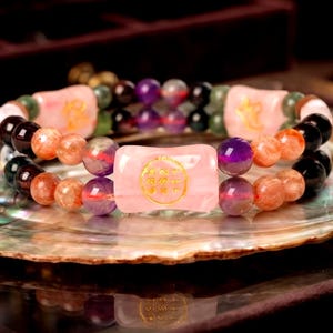 May include: A multi-strand beaded bracelet featuring polished round beads in shades of orange, purple, and black. The bracelet includes two rectangular pink stone beads, each engraved with gold-colored Chinese characters, arranged symmetrically.