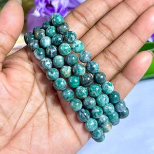 May include: A beaded bracelet featuring round, polished beads in shades of green and gray. The beads are strung together, creating a flexible band. The bracelet is held in a hand, showcasing the natural variations in the stone.