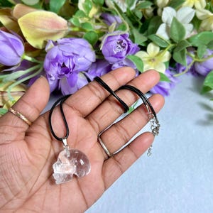 May include: A clear quartz skull pendant on a black cord necklace. The pendant is held in a hand, with a floral background of purple and white flowers. The necklace has a silver-colored clasp.