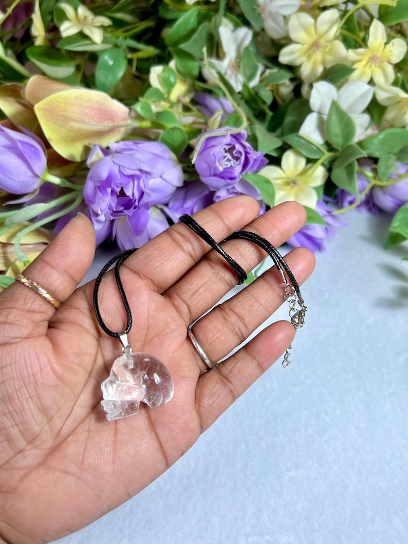 May include: A clear quartz skull pendant on a black cord necklace. The pendant is held in a hand, with a floral background of purple and white flowers. The necklace has a silver-colored clasp.