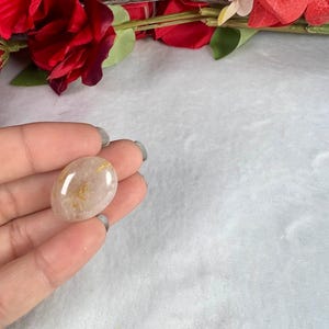 May include: A polished, oval-shaped gemstone with a translucent, light brown hue and golden inclusions. The stone is held in a hand, with a soft, white background and red floral accents in the upper portion of the image.
