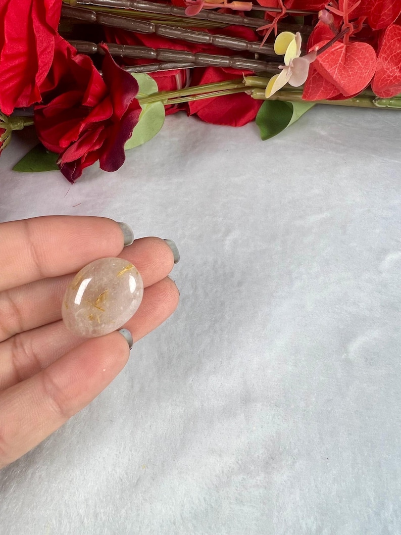 May include: A polished, oval-shaped gemstone with a translucent, light brown hue and golden inclusions. The stone is held in a hand, with a soft, white background and red floral accents in the upper portion of the image.