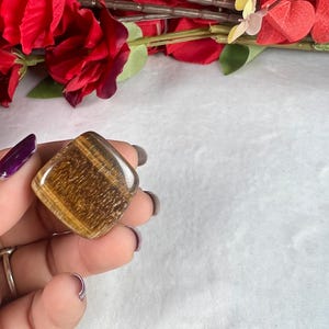 May include: A polished, square-shaped tiger's eye gemstone, displaying bands of golden brown and amber hues. The stone is held against a backdrop of red flowers and a white surface.