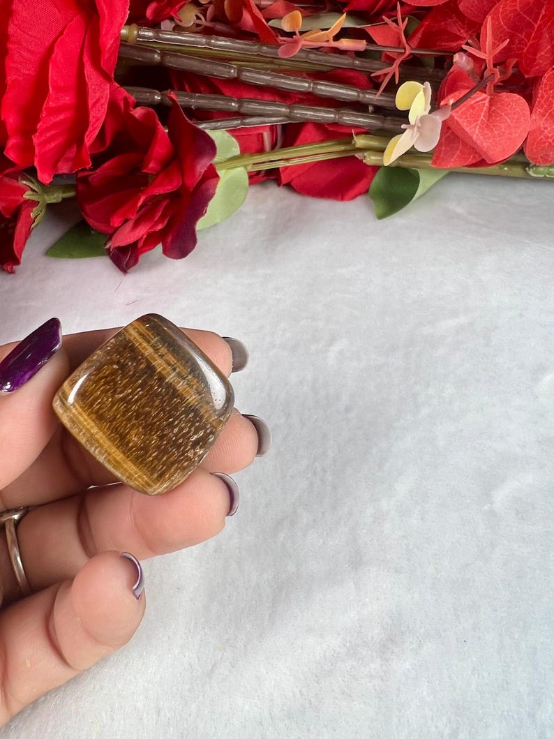 May include: A polished, square-shaped tiger's eye gemstone, displaying bands of golden brown and amber hues. The stone is held against a backdrop of red flowers and a white surface.