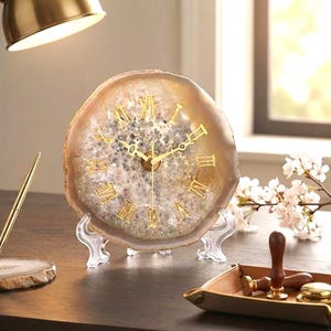 May include: A round clock made from a slice of natural agate stone with a translucent, crystalline center and gold-colored Roman numeral markers. The clock features gold-colored hands and is displayed on a clear plastic stand.