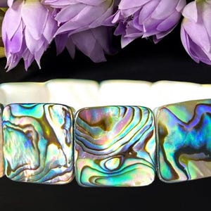 May include: A stretch bracelet composed of square-shaped abalone shell tiles. Each tile displays a natural iridescent pattern with swirling hues of blue, green, and purple. The interior of the bracelet is white, and the tiles are linked together.