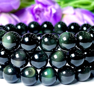 May include: A layered bracelet made of polished, round, black obsidian beads. The beads have a reflective, glossy surface with hints of green and gold. The background features blurred purple flowers and green foliage.