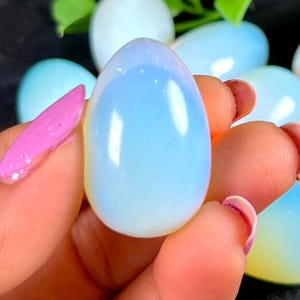 Puede incluir: Una piedra preciosa pulida de forma ovalada, con un aspecto opalescente y nacarado, sostenida en una mano. La piedra presenta un degradado de tonos azul claro y blanco. Otras piedras similares son visibles en el fondo.