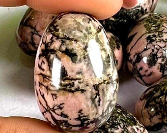 May include: Polished, oval-shaped gemstones with a pink base color and black veining. The stones have a glossy finish and are held by a person. Other similar stones are in the background.