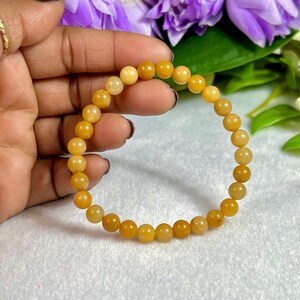 May include: A beaded bracelet featuring small, round, yellow-orange beads. The bracelet is held against a backdrop of purple flowers and green leaves. The beads appear to be strung on an elastic cord, forming a complete circle.