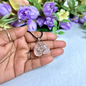 May include: A clear quartz crystal skull pendant on a black cord necklace. The skull is intricately carved and held in a hand, with a blurred floral background of purple and white flowers. The pendant is a unique piece of jewelry.