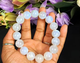 Könnte beinhalten: Ein Armband aus glatten, runden, durchscheinenden weißen Mondsteinperlen mit einem dezenten irisierenden blauen Schimmer. Die Perlen sind kreisförmig aufgereiht und weisen eine polierte Oberfläche auf, die das Licht auf der gesamten Fläche reflektiert.