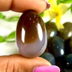 May include: A polished, egg-shaped stone with a gradient of brown to light purple hues. The smooth surface reflects light, and the stone is held between two fingers. Other similar stones are visible in the background.