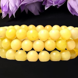 May include: A multi-strand bracelet composed of three rows of smooth, round, pale yellow beads. The beads have a consistent, opaque, and matte finish, arranged in a circular formation to create a layered appearance on a dark background.
