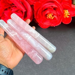 May include: Three rose quartz crystal massage wands in varying shades of pink and white are held in a hand. The wands are cylindrical and smooth, with a polished finish. Red roses are in the background.