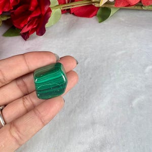 May include: A polished malachite stone with dark and light green bands. The stone is held in a hand, with red flowers in the background. The malachite is a square shape, and the green bands create a striped pattern.