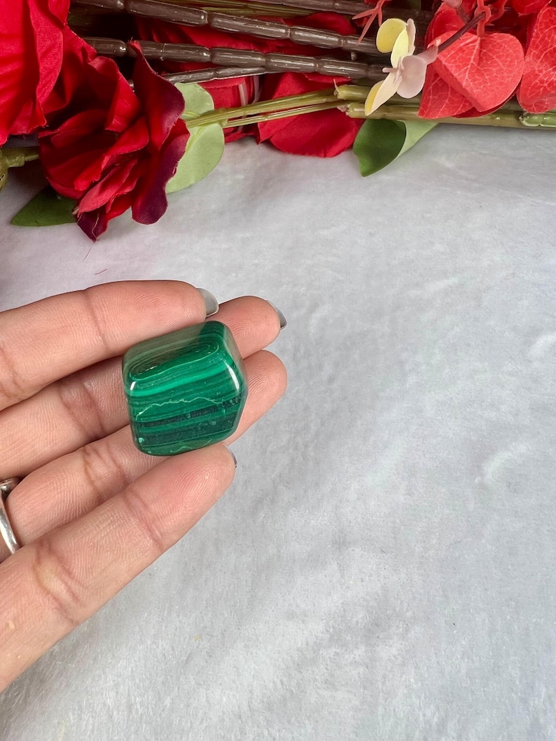 May include: A polished malachite stone with dark and light green bands. The stone is held in a hand, with red flowers in the background. The malachite is a square shape, and the green bands create a striped pattern.