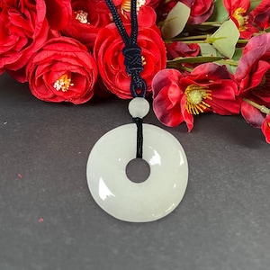 May include: A white jade pendant with a circular design and a central hole, suspended from a black cord. The pendant is smooth and polished, with a subtle sheen. The background features red roses.