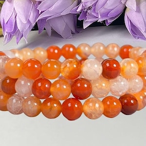 Peut inclure: Un bracelet multi-rangs composé de perles rondes et polies dans des tons variés d'orange, de rouge et de blanc translucide. Les perles présentent des craquelures internes naturelles et des dégradés de couleurs, disposées en cercle.