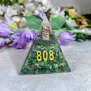 May include: A pyramid-shaped decorative object with a clear top and green base. The base is filled with green stones and the gold numbers "808" are visible. A clear crystal is wrapped with copper wire at the top.