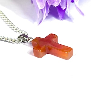 May include: A silver chain necklace with a polished, orange-red cross pendant. The cross is made of a translucent material, and the chain is a classic curb style. The background is white with a hint of purple.