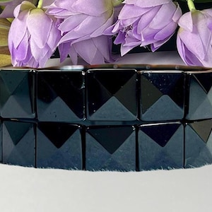 May include: A black bracelet featuring a series of square, pyramid-shaped studs arranged in a single row. The bracelet has a glossy, reflective surface and is positioned horizontally, showing a clear reflection of the studs on the surface below.