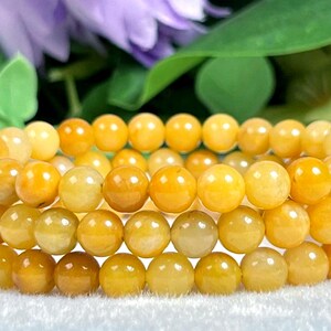 May include: Three bracelets of round, yellow-orange beads. The beads have a smooth, polished appearance and vary slightly in color. The bracelets are stacked, with a blurred background of green leaves and purple flowers.